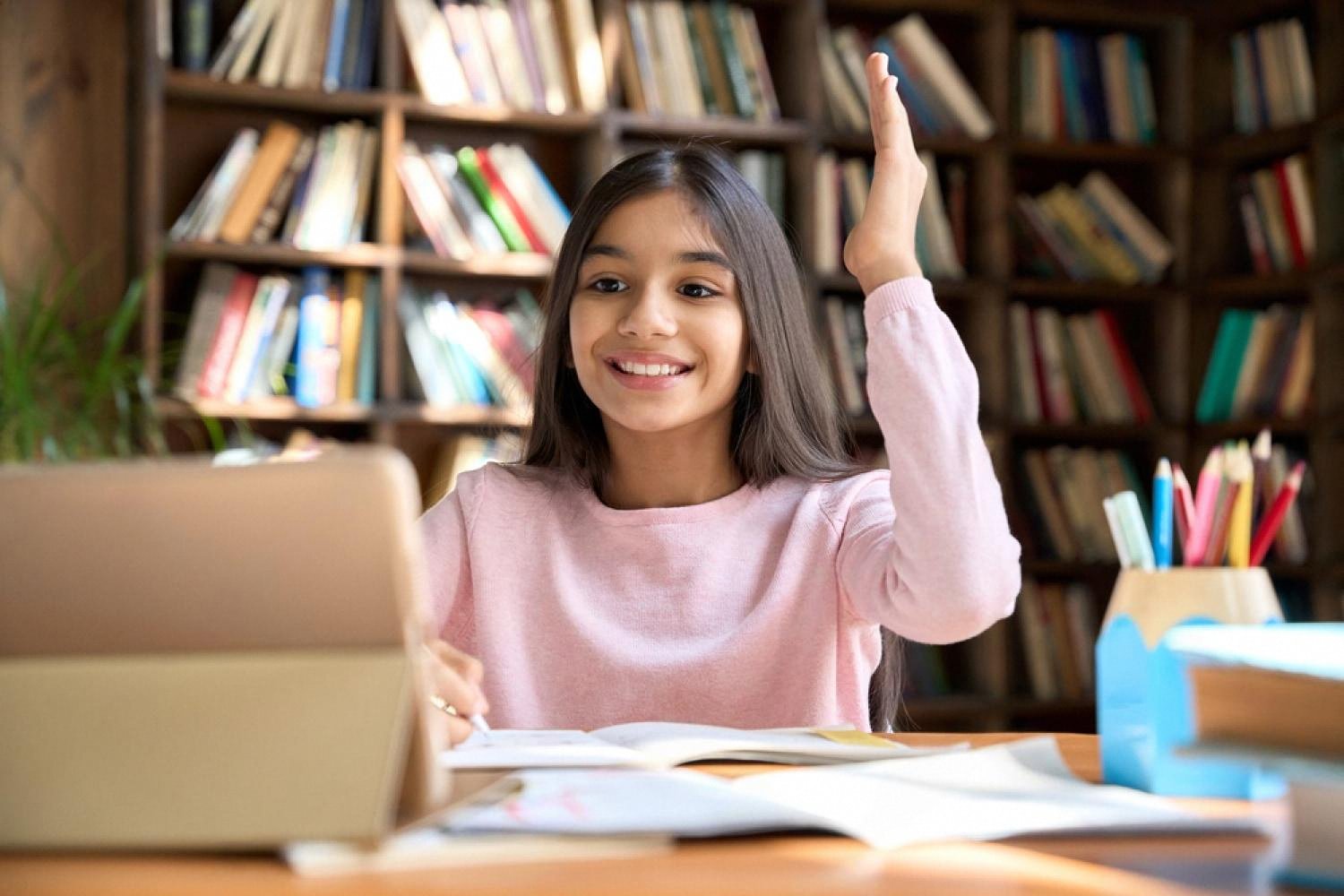 Smiling schoolgirl raising her hand while studying at a desk with books and a tablet